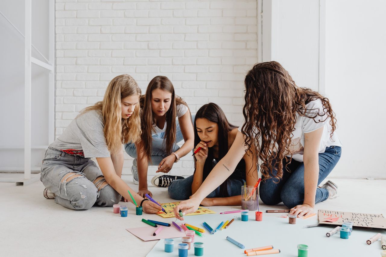 Home Four teenage girls collaboratively painting and drawing on a floor with bright colors, embracing creativity.